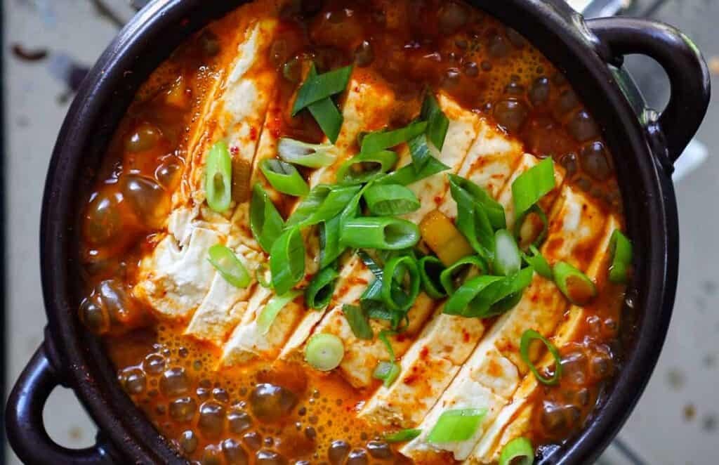 BEST Doenjang Jjigae (된장찌개) - Spicy and Delicious. 2 A bowl of bubbling doenjang jjigae is topped with sliced tofu and garnished with chopped green onions. The stew appears to have a rich, spicy red broth, served in a dark, circular pot.