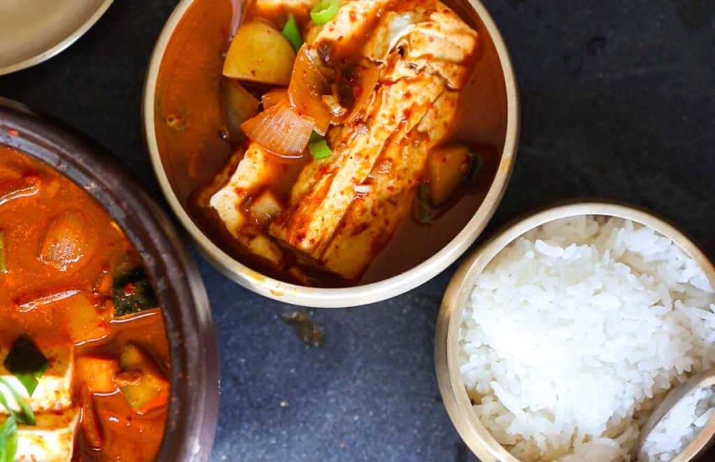 BEST Doenjang Jjigae (된장찌개) - Spicy and Delicious. 3 A bowl of spicy doenjang jjigae with tofu, zucchini, onions, and other vegetables is next to a bowl of white rice on a dark surface. Another partial bowl of stew is visible, suggesting a Korean meal setting.