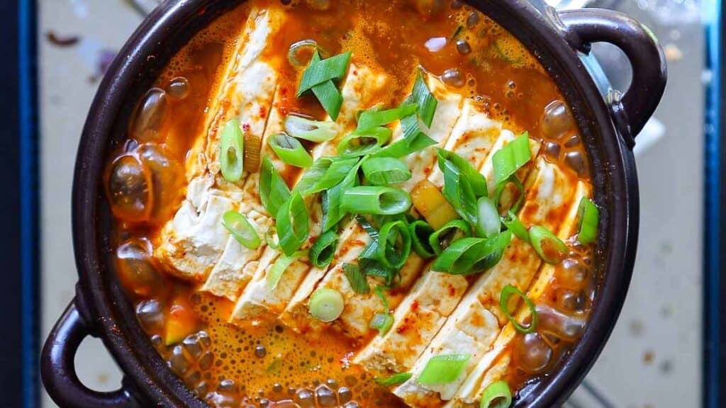 BEST Doenjang Jjigae (된장찌개) - Spicy and Delicious. 1 A bowl of spicy doenjang jjigae with a rich orange broth, topped with sliced green onions. The tofu is cut into rectangular pieces and arranged neatly in the soup, which is served in a dark ceramic bowl.
