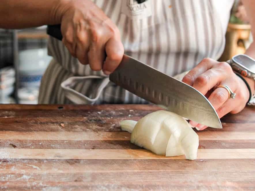 Creamy Cozy Kale White Bean Soup (EASY & Vegan). 9 A person wearing a striped apron uses a large kitchen knife to slice a white onion on a wooden cutting board. Their left hand holds the onion steady while their right hand grips the knife. They are also wearing a watch on their left wrist.