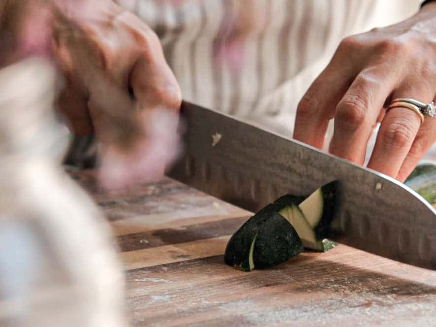 BEST Doenjang Jjigae (된장찌개) - Spicy and Delicious. 7 Close-up of hands slicing a zucchini on a wooden cutting board. One hand holds the zucchini steady while the other uses a knife to cut. The person is wearing a ring on their left hand.