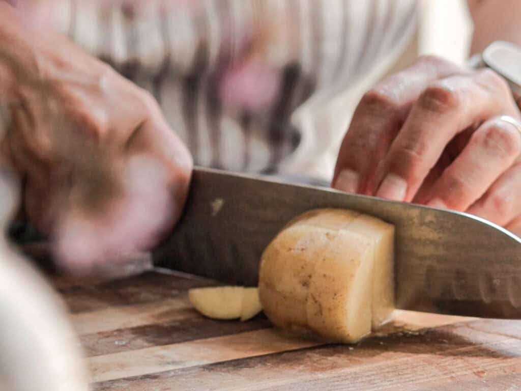 Creamy Cozy Kale White Bean Soup (EASY & Vegan). 8 Close-up of hands slicing a potato with a large knife on a wooden cutting board. The person is wearing a striped apron, and the focus is on the knife and potato. The background is blurred.