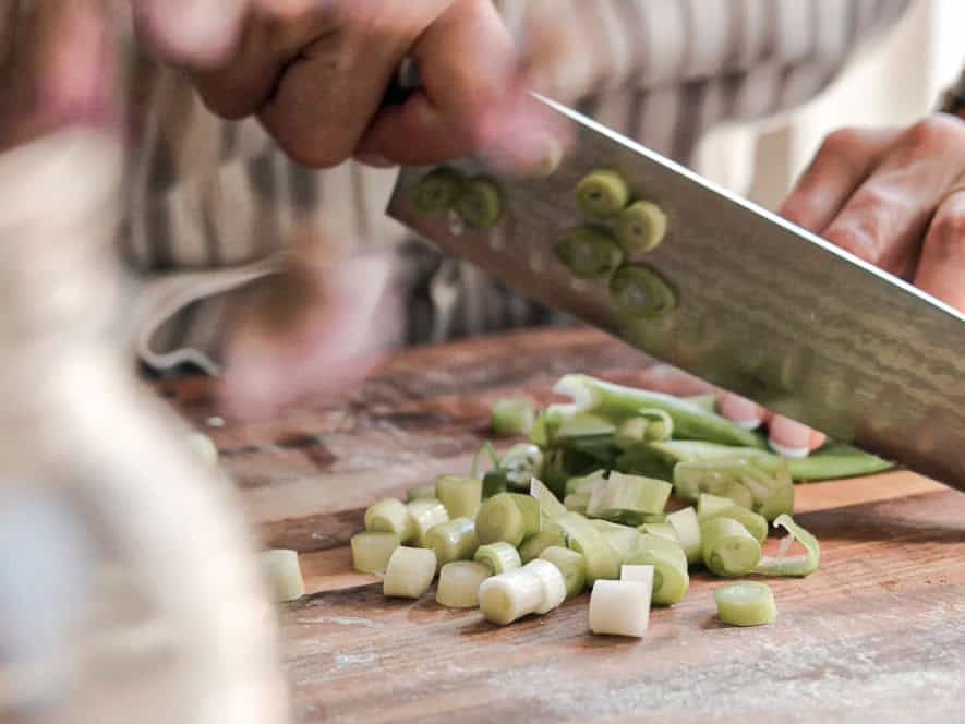 BEST Doenjang Jjigae (된장찌개) - Spicy and Delicious. 9 A person chopping green onions on a wooden cutting board. The focus is on the hands and the knife, with sliced green onion pieces scattered around.