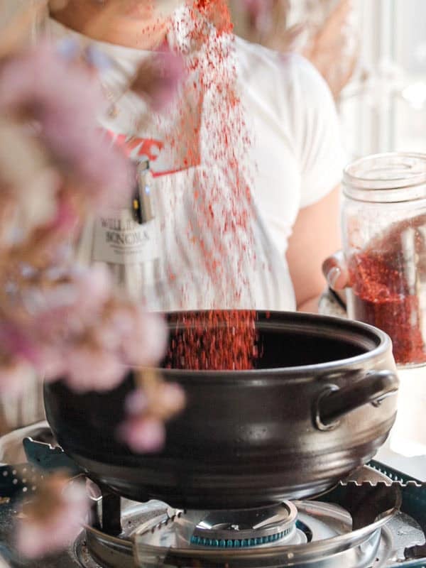 BEST Doenjang Jjigae (된장찌개) - Spicy and Delicious. 5 Person pouring gochugaru from a jar into a black pot on a gas stove. The pot has handles, and the person is wearing a white shirt. Blurred foreground elements include pinkish flowers.