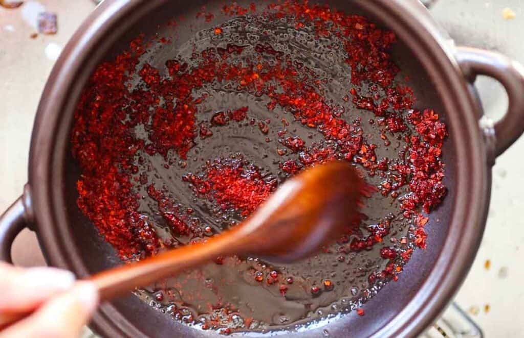 BEST Doenjang Jjigae (된장찌개) - Spicy and Delicious. 14 A hand is stirring gochugaru in a black pot with a wooden spoon. The paste is sizzling and spreading across the surface of the pot, creating a vibrant red swirl against the dark background.
