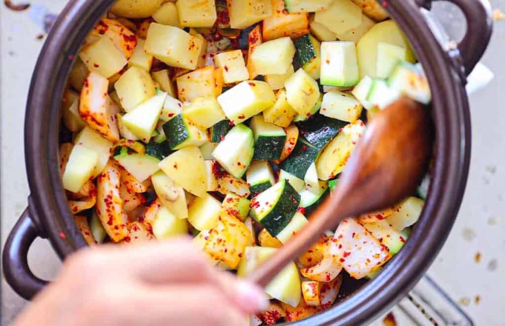 BEST Doenjang Jjigae (된장찌개) - Spicy and Delicious. 15 A pot filled with chopped potatoes and zucchini seasoned with red pepper flakes. A hand is stirring the mixture with a wooden spoon. The vegetables are vibrant, with green and yellow hues, contrasting with the red spices.