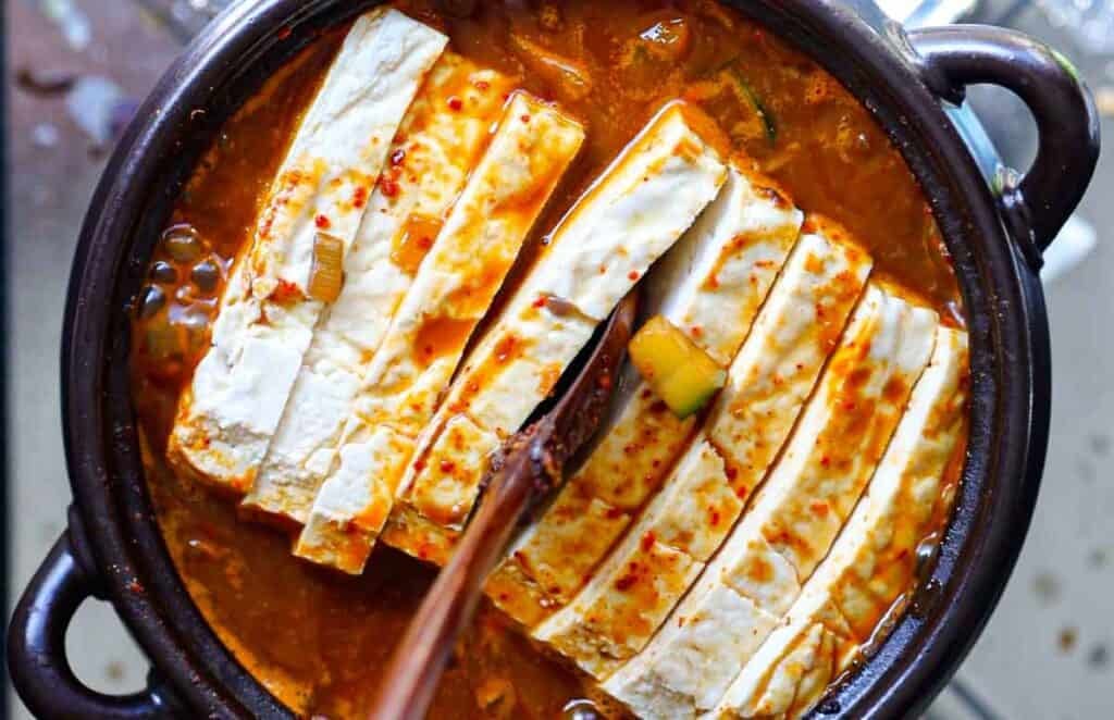 BEST Doenjang Jjigae (된장찌개) - Spicy and Delicious. 20 Close-up of a traditional Korean stew in a black pot, featuring thick slices of tofu immersed in a rich, spicy red broth. The soup contains visible ingredients such as sliced leeks and chili flakes. Steam rises, indicating its hot and freshly made.