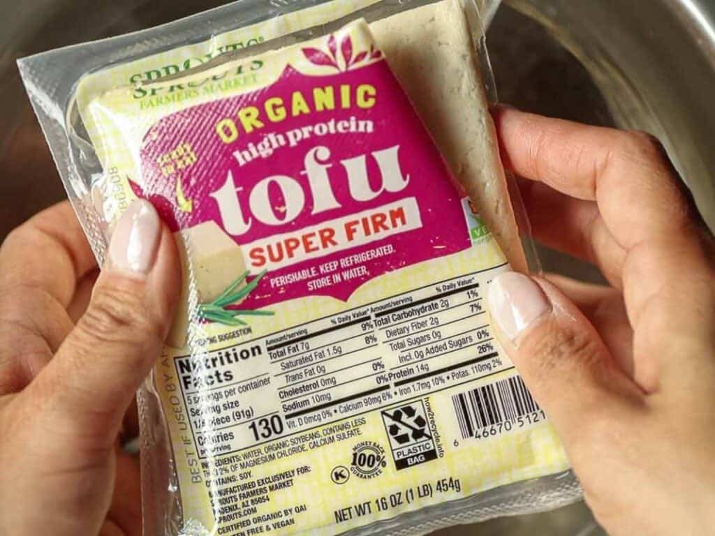 Delicious and Easy Vegan Cashew Nut Stir Fry Recipe. 4 Close-up of hands holding a package of organic, high-protein, super firm tofu. The label shows it as non-GMO, with nutrition facts visible. The package weighs 16 oz (454g).