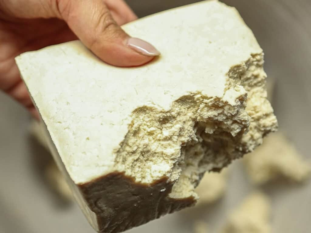 Delicious and Easy Vegan Cashew Nut Stir Fry Recipe. 13 A hand holding a block of super firm tofu with pieces crumbling off. The tofu has a textured surface and a light beige color, with visible areas where chunks have broken away.