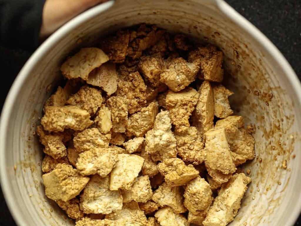 Delicious and Easy Vegan Cashew Nut Stir Fry Recipe. 14 A large bowl filled with chunks of super firm tofu marinating in a brown sauce. The tofu pieces are irregular in shape, and the marinade adds a textured, glazed appearance. One hand is partially visible holding the bowl.