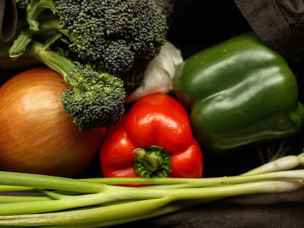 Delicious and Easy Vegan Cashew Nut Stir Fry Recipe. 6 A collection of fresh vegetables: a bunch of broccoli, a green bell pepper, a red bell pepper, an onion, and green onions. They are arranged together on a dark background.