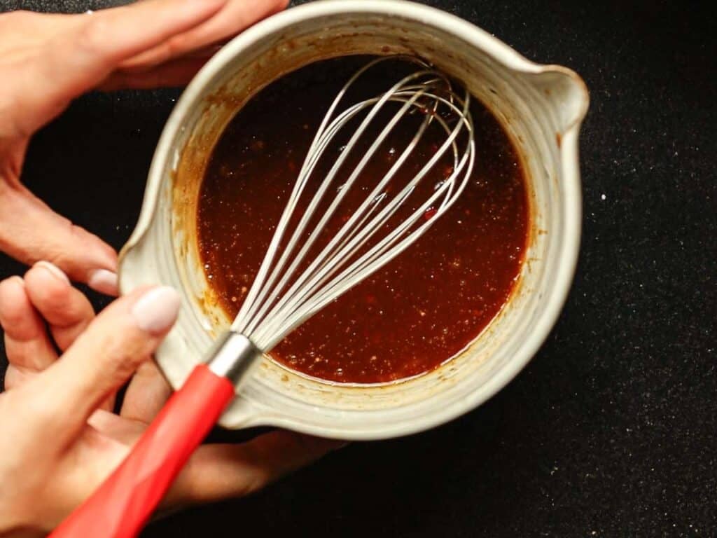 Delicious and Easy Vegan Cashew Nut Stir Fry Recipe. 18 Hands holding a green bowl with reddish-brown sauce being whisked by a red-handled whisk against a black background.