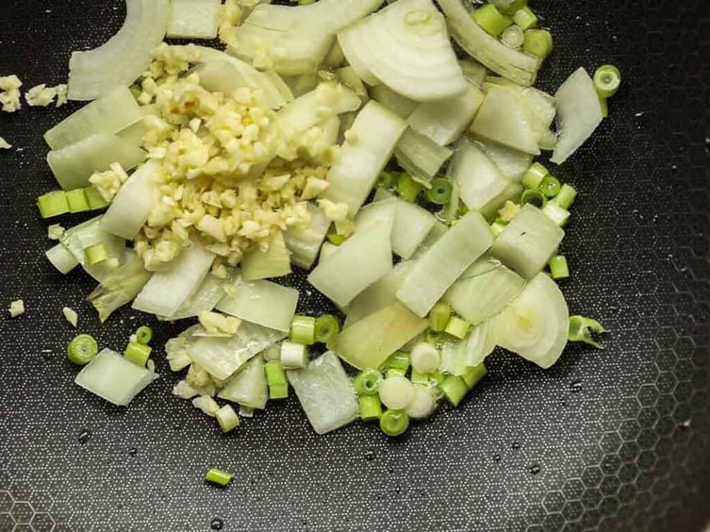 Delicious and Easy Vegan Cashew Nut Stir Fry Recipe. 19 Chopped onions, garlic, and green onions are scattered on a hexagon-textured dark surface, ready for cooking.