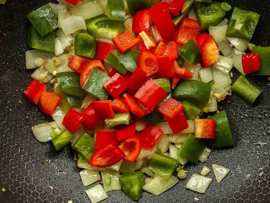 Delicious and Easy Vegan Cashew Nut Stir Fry Recipe. 20 Chopped red and green bell peppers and onions are being sautéed in a pan. The vegetables are vibrant and colorful, and the pan has a textured non-stick surface.