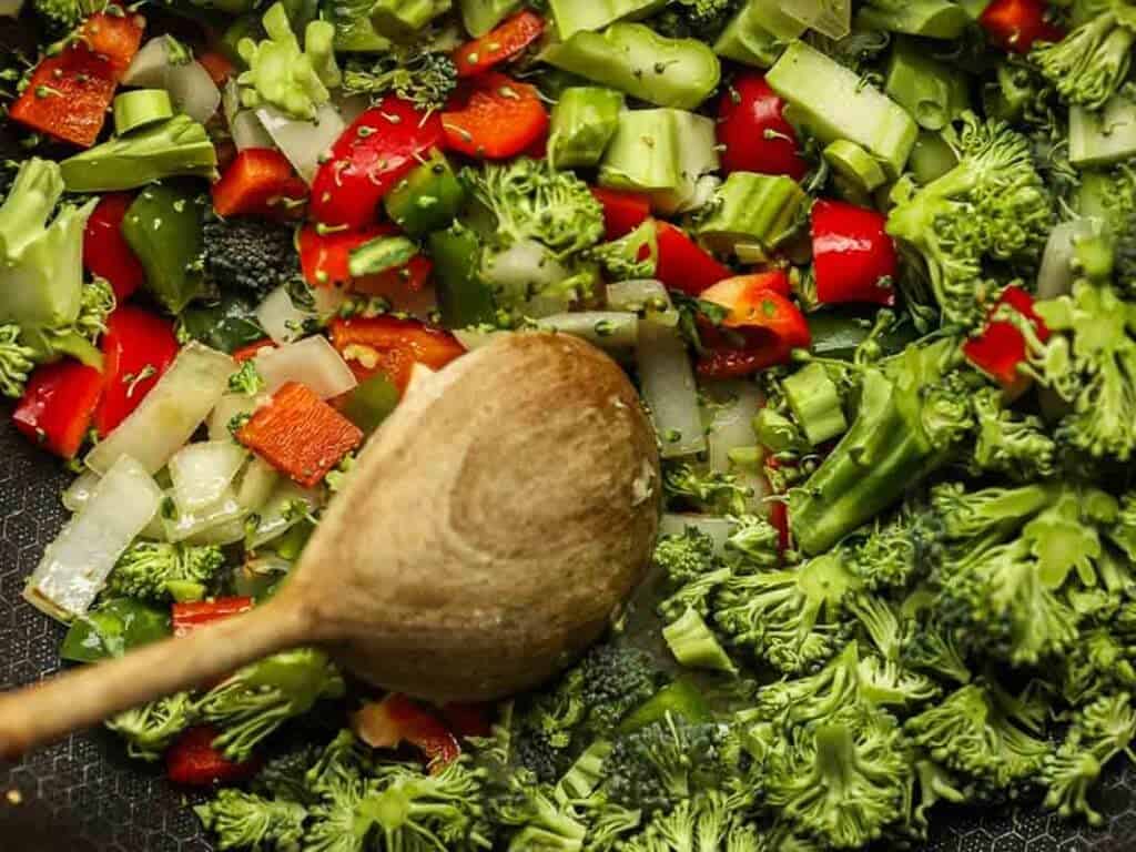 Delicious and Easy Vegan Cashew Nut Stir Fry Recipe. 21 Close-up of chopped broccoli, red bell peppers, onions, and small green vegetables in a pan. A wooden spoon is stirring the colorful mix, suggesting a healthy vegetable stir-fry being prepared.