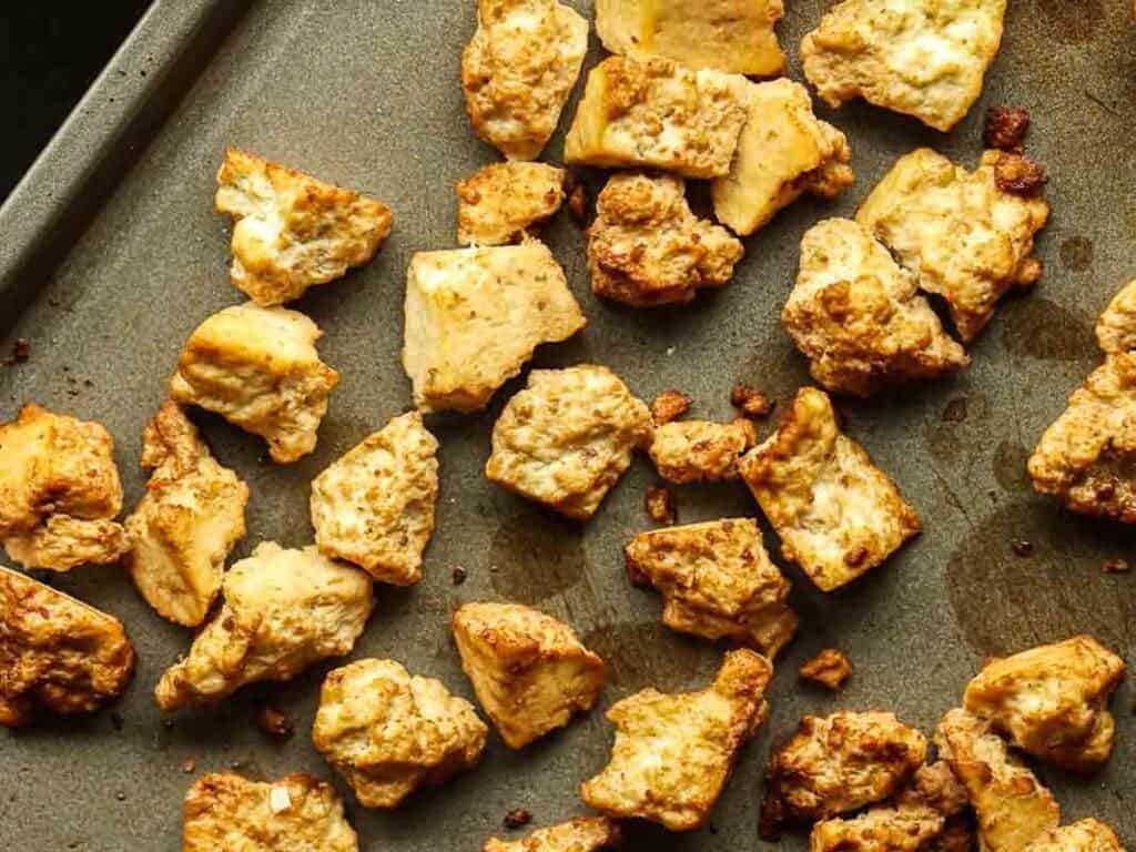 Delicious and Easy Vegan Cashew Nut Stir Fry Recipe. 17 Golden-brown roasted tofu pieces scattered on a dark baking sheet, displaying a crispy texture and lightly charred edges.