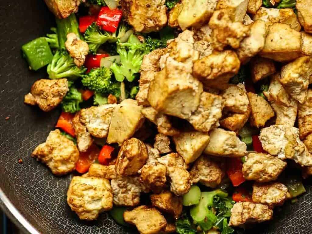 Delicious and Easy Vegan Cashew Nut Stir Fry Recipe. 22 Stir-fried tofu cubes with broccoli, red bell peppers, and other vegetables in a frying pan. The tofu is golden brown, while the vegetables are vibrant and fresh.