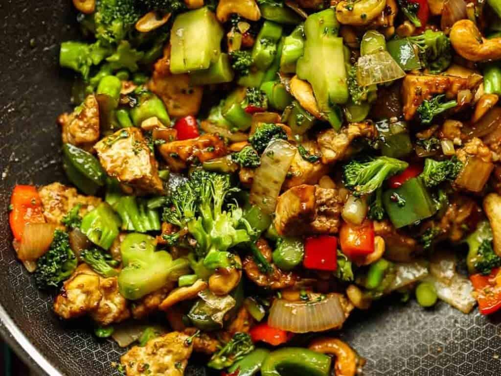 Delicious and Easy Vegan Cashew Nut Stir Fry Recipe. 3 Close-up of a stir-fry featuring a mix of vibrant vegetables including broccoli, bell peppers, and onions, along with tofu and cashews, all seasoned and cooked in a skillet.