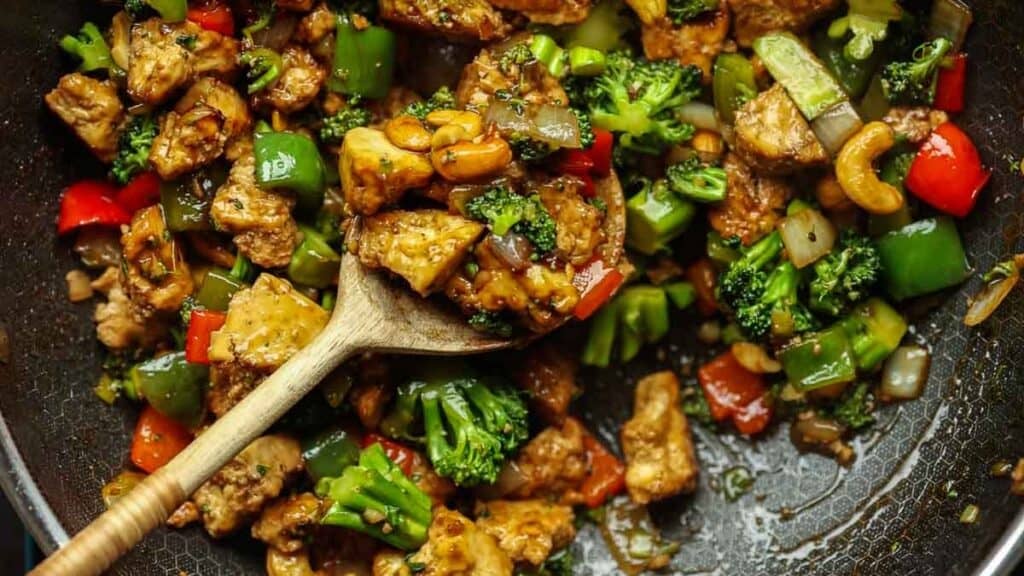 Delicious and Easy Vegan Cashew Nut Stir Fry Recipe. 1 A wooden spoon holds a cashew nut stir fry with tofu, broccoli, green and red bell peppers, cashews, and onions in a pan. The dish is colorful and appears to be seasoned with a glossy sauce.
