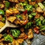 30+ AMAZING VEGAN Holiday Recipes Everyone's gonna Love. 13 A close-up of cashew nut stir fry with tofu and vegetables in a pan. The dish includes broccoli, bell peppers, and peanuts, all tossed in a savory sauce. A wooden spoon holds a portion of the mix, highlighting the vibrant colors and texture.