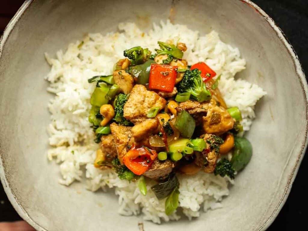 Delicious and Easy Vegan Cashew Nut Stir Fry Recipe. 2 A bowl of white rice topped with a colorful cashew nut stir fry featuring pieces of tofu, red and green bell peppers, broccoli, and cashews.