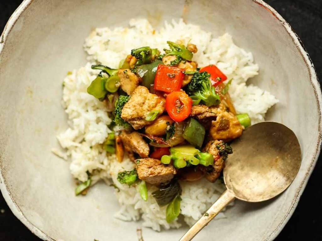 Delicious and Easy Vegan Cashew Nut Stir Fry Recipe. 25 A white bowl filled with white rice topped with a colorful cashew nut stir fry including broccoli, red peppers, and pieces of tofu. A bronze spoon rests on the side of the bowl.