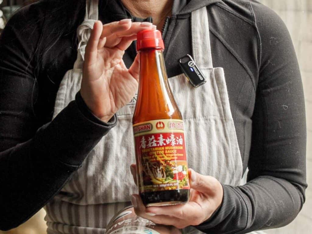 Delicious and Easy Vegan Cashew Nut Stir Fry Recipe. 10 A person wearing a black shirt and striped apron holds a bottle labeled Vegetarian Mushroom Stir-Fry Sauce with red accents. The person’s face is not visible.