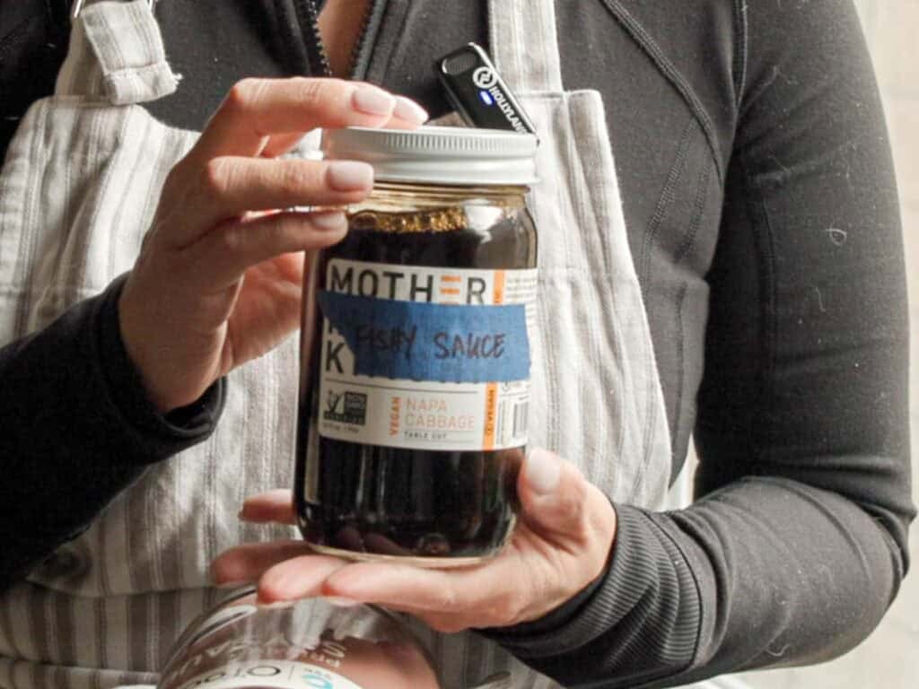 BEST Baby Bok Choy Kimchi Recipe - Easy & Delicious 13 Person holding a jar labeled Fishy Sauce with โMotherโ and โNapa Cabbageโ on a sticker. They are wearing a black top and striped apron. Another jar is partially visible at the bottom.