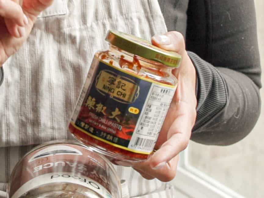 Delicious and Easy Vegan Cashew Nut Stir Fry Recipe. 12 A person wearing an apron holds a jar of Ning Chi brand chili paste in one hand and another jar with an obscured label in the other.