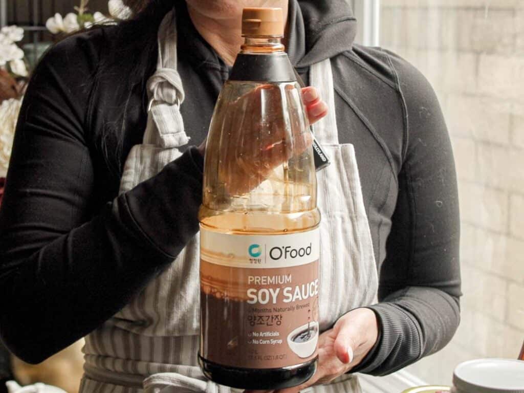 Delicious and Easy Vegan Cashew Nut Stir Fry Recipe. 8 A person in a gray apron is holding a large bottle of OFood Premium Soy Sauce. The label includes text in Korean and English. The person is standing near a window with a neutral background.
