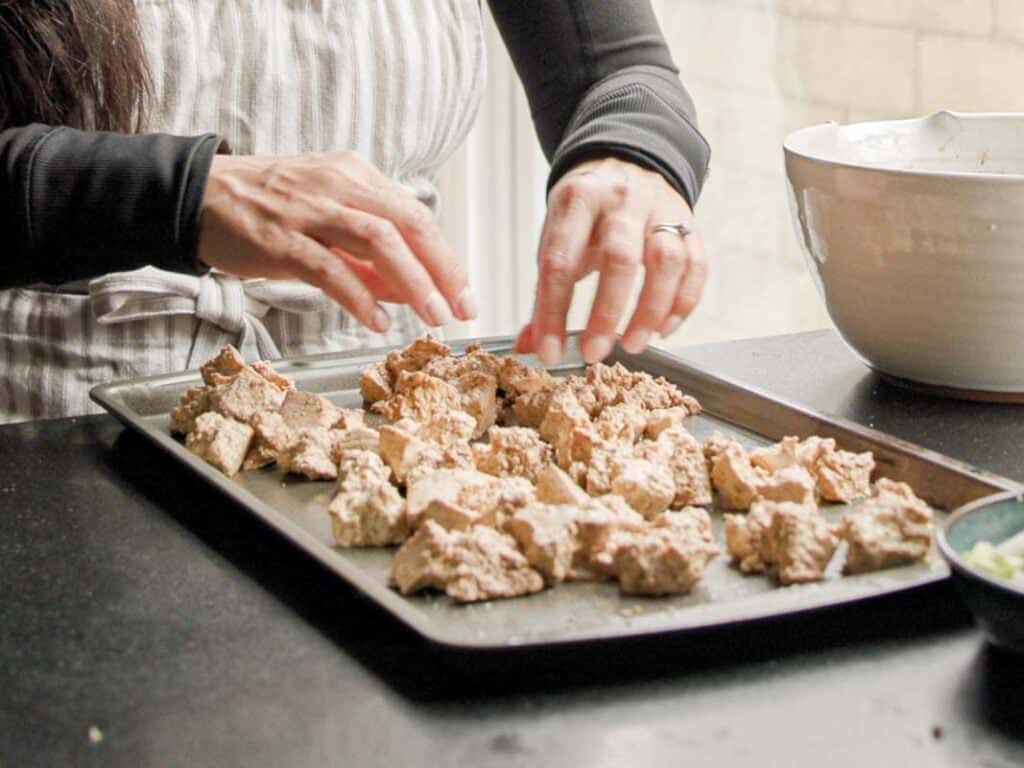 Delicious and Easy Vegan Cashew Nut Stir Fry Recipe. 15 A person wearing a striped apron arranges pieces of textured, tofu on a baking tray. There is a large white bowl nearby. The scene suggests preparation for baking or roasting.