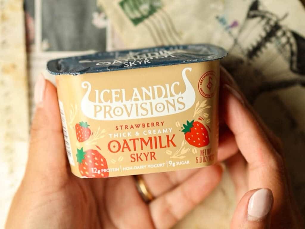 Viral Dubai Chocolate Bar with Strawberry Filling (Vegan). 8 Person holding a container of Icelandic Provisions Strawberry Oatmilk Skyr. The packaging is tan with red and blue accents, featuring strawberries and nutritional details.