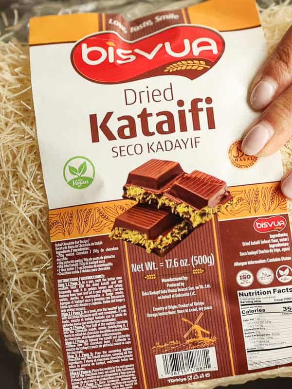 Viral Dubai Chocolate Bar with Strawberry Filling (Vegan). 4 A hand holding a package of Bisvua Dried Kataifi. The package shows images of chocolate-covered kataifi. It features a green vegan label and nutritional information. The background contains strands of kataifi pastry.