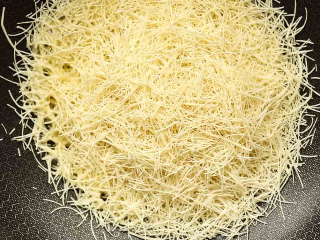 A close-up of dried kataifi in a pan. The pasta consists of thin, short strands arranged in a circular pattern on a dark, textured surface.