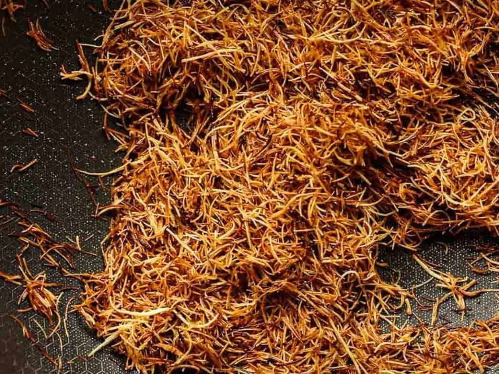 Close-up of a pile of toasted kataifi with a brownish-golden hue, spread out on a dark surface. The thin, stringy fibers are densely packed, showcasing their intricate and delicate texture.