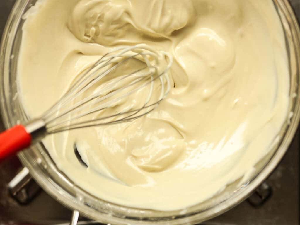 Viral Dubai Chocolate Bar with Strawberry Filling (Vegan). 13 A whisk in a glass bowl filled with smooth, melted white chocolate. The mixture is light-colored and evenly blended, with the whisk partially submerged and visible from a top view.
