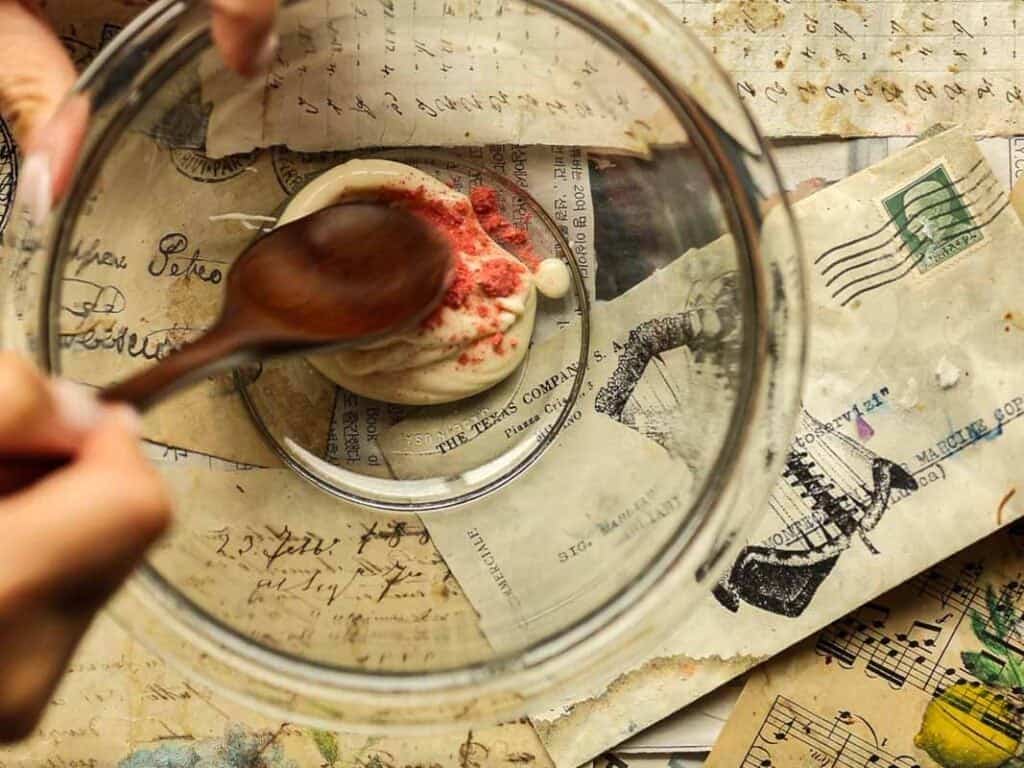 A person stirs white chocolate with red sprinkles in a glass bowl using a wooden spoon. The background is scattered with vintage papers, handwritten notes, stamps, and musical scores.