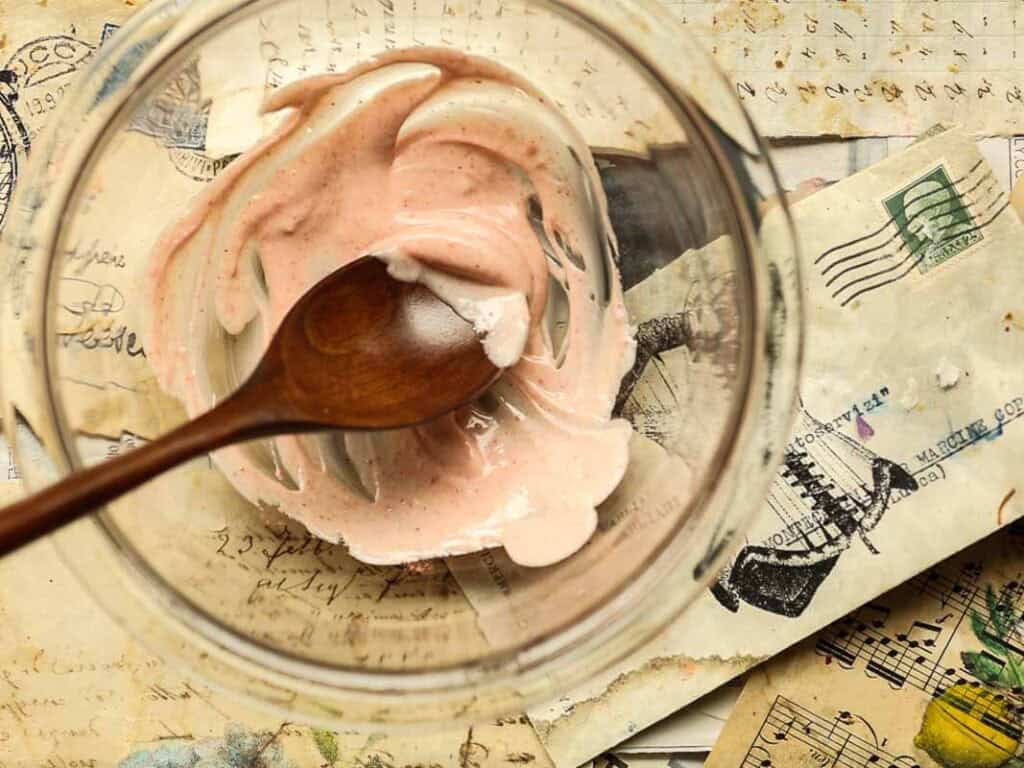 A glass bowl with remnants of swirled white chocolate and freeze dried strawberry grounds and a wooden spoon rests on top of scattered vintage letters and postcards.
