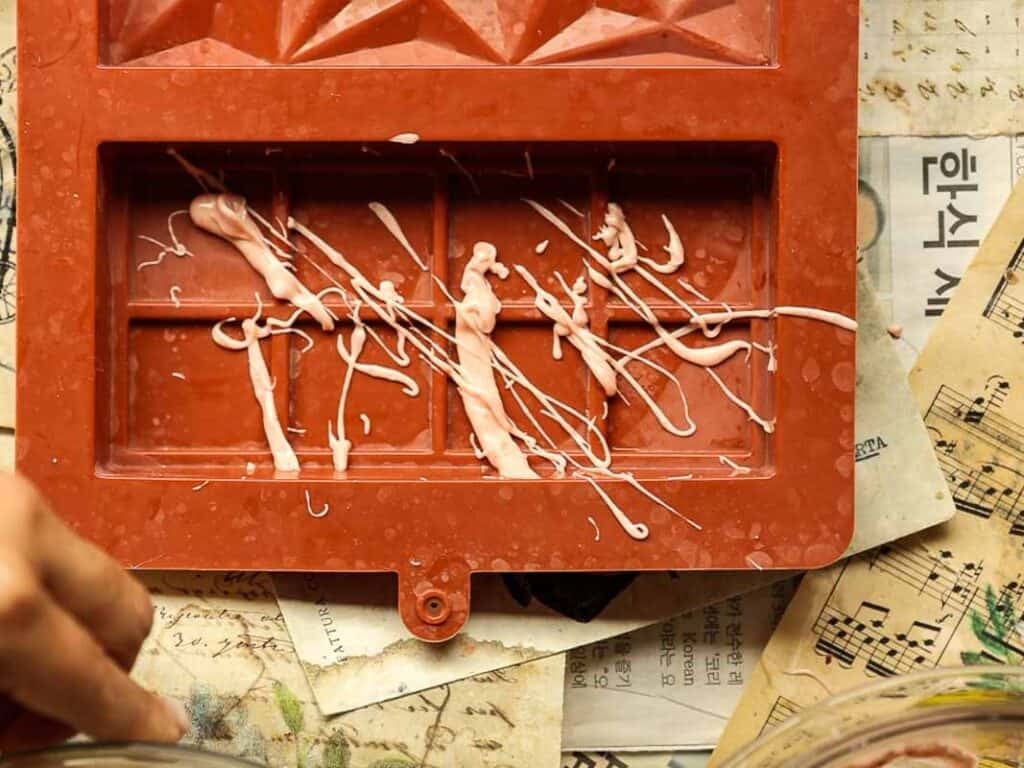 A red chocolate mold with white chocolate drizzles inside rests on a cluttered surface covered with Korean text, sheet music, and vintage paper. Part of a hand is visible in the lower left corner.