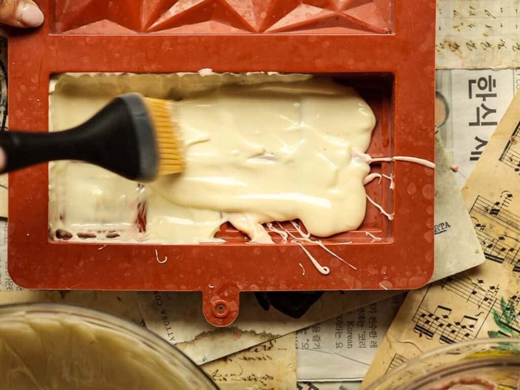 A person uses a brush to spread white chocolate in a red, triangular-patterned mold. Beneath the mold are sheets of newspaper with text and musical notes.