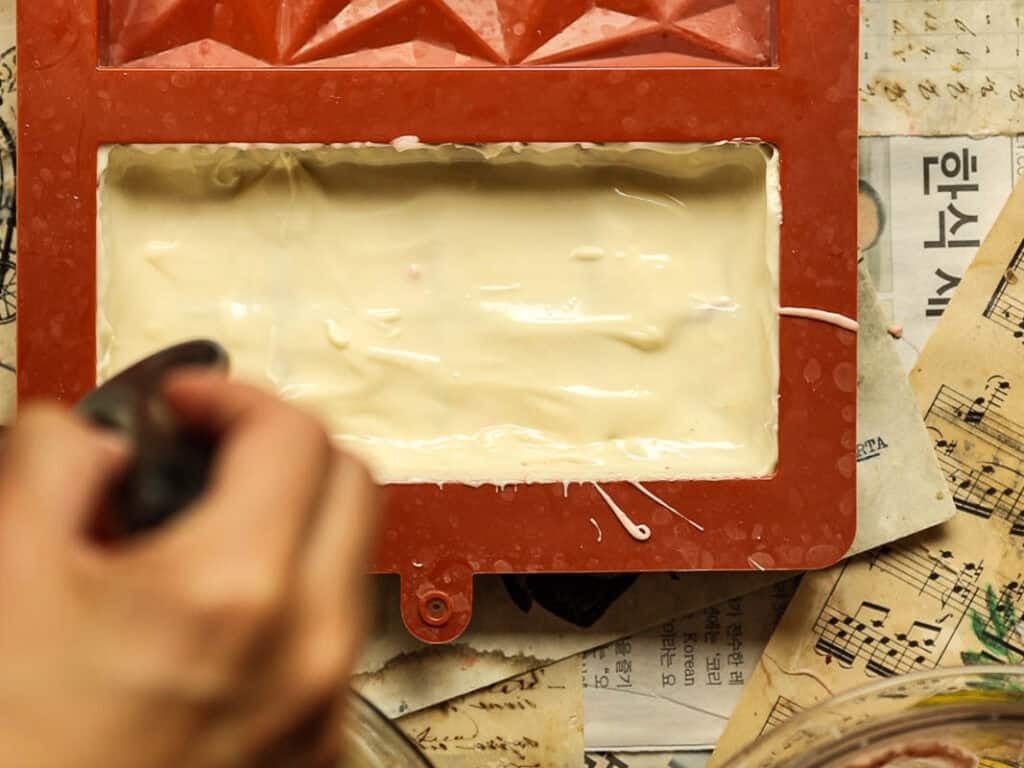 A person is pouring a melted white chocolate into a rectangular red silicone mold placed on sheets of old newspaper and music paper. The mold is filled with a light-colored batter.