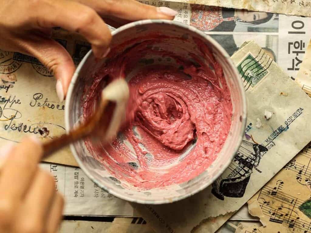 Viral Dubai Chocolate Bar with Strawberry Filling (Vegan). 19 A person mixes pink mixture in a bowl using a brush. The bowl is placed on sheets of newspaper with text in different languages. The setting looks like an art or craft workspace.