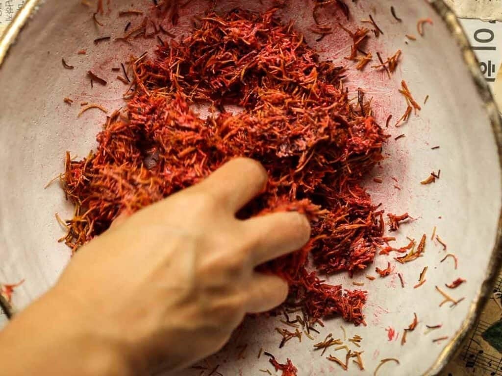 Viral Dubai Chocolate Bar with Strawberry Filling (Vegan). 20 A hand mixing reddish kataifi threads in a white ceramic bowl. The strands are thin and have a vibrant hue, contrasting with the bowls interior. The action suggests preparation or examination of the spice.