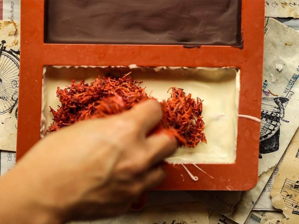 A hand is placing shredded kataifi into a rectangular mold filled with white and dark chocolate. The mold is placed on printed paper, and there are smudges of chocolate around the area.