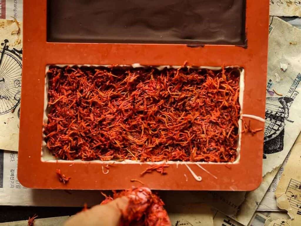 A tray filled with bright red kataifi strands on top of a background with scattered illustrations. The trays border is orange, and part of a persons hand is visible at the bottom, holding some saffron.