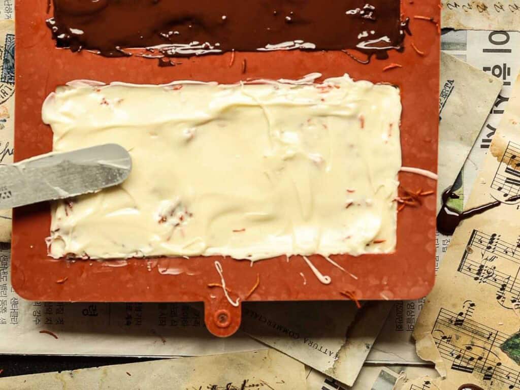A red silicone mold holds partially solidified dark and white chocolate layers. A spatula rests on the white chocolate, with scattered shredded kataifi. The background features layered sheet music and pages with foreign text.