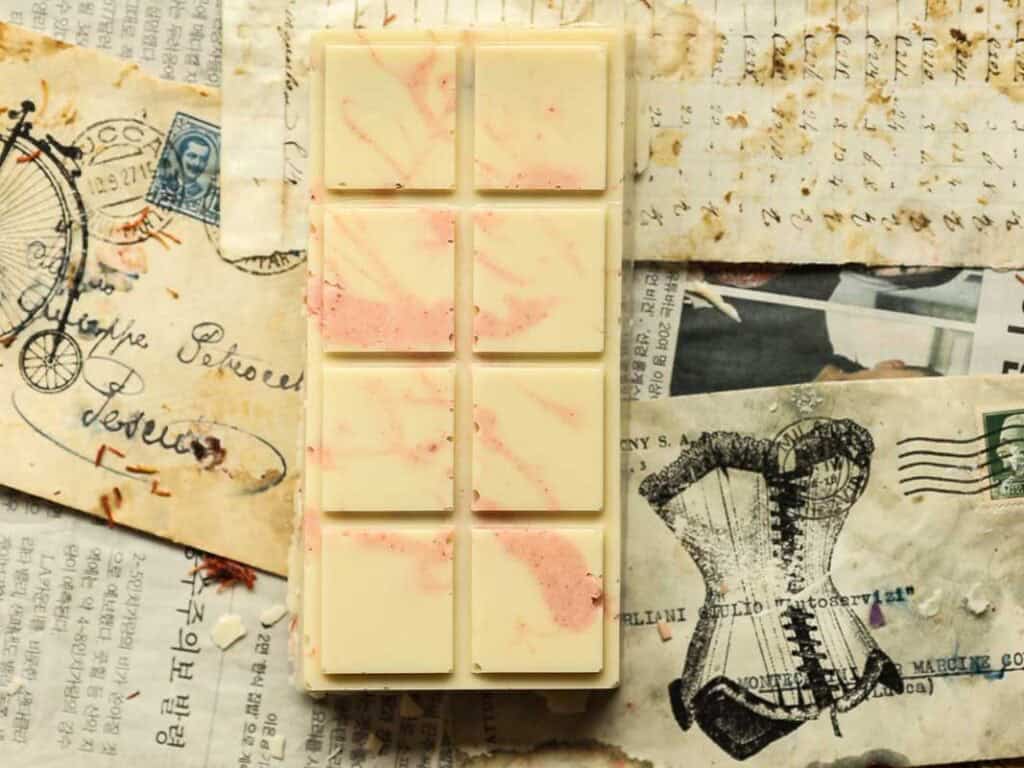 Viral Dubai Chocolate Bar with Strawberry Filling (Vegan). 25 A white chocolate bar with pink swirls rests on a background of vintage paper featuring old bicycle and corset illustrations, postage stamps, and handwritten text.