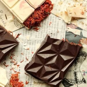 Viral Dubai Chocolate Bar with Strawberry Filling (Vegan). 26 Close-up of geometric dark and white dubai chocolate bars with kataifi scattered around. The chocolates rest on vintage newspaper pieces, with a partially bitten piece showing texture.
