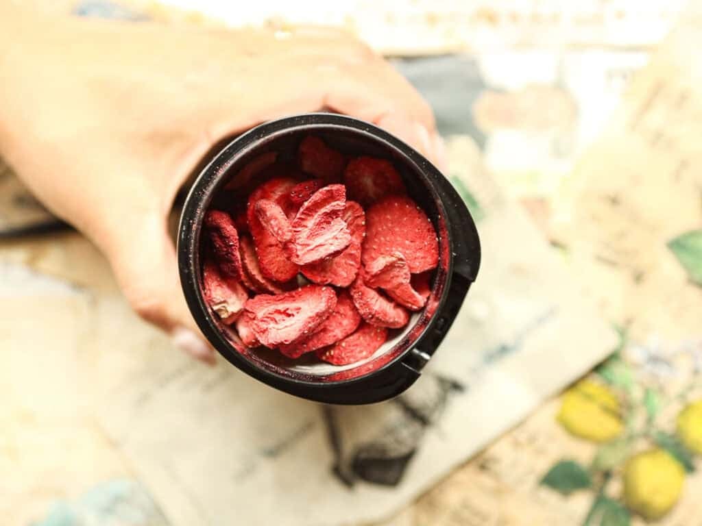 A hand holds a black cup filled with freeze-dried strawberry slices. The background displays vintage-style paper sheets with text and illustrations, creating a nostalgic atmosphere.