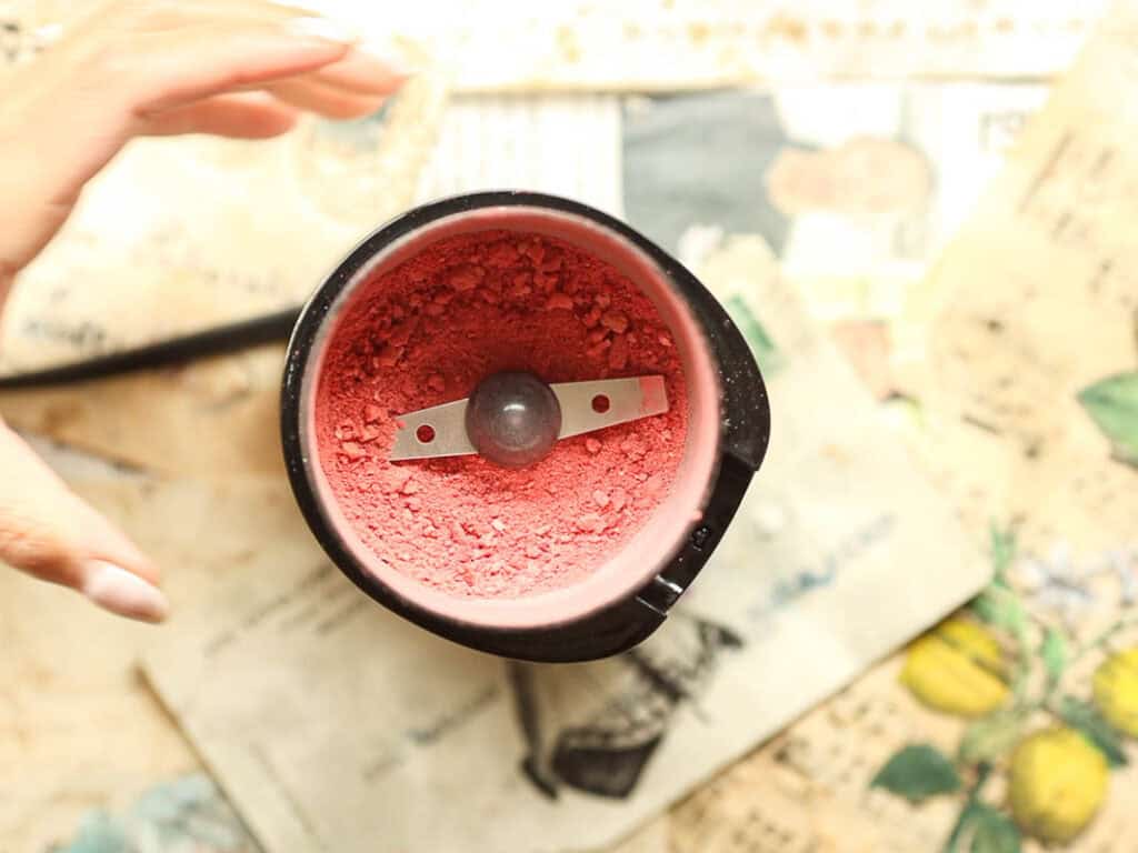 Top view of a spice grinder containing fine pink powder. A hand reaches toward it. Background features various papers with illustrations and text.